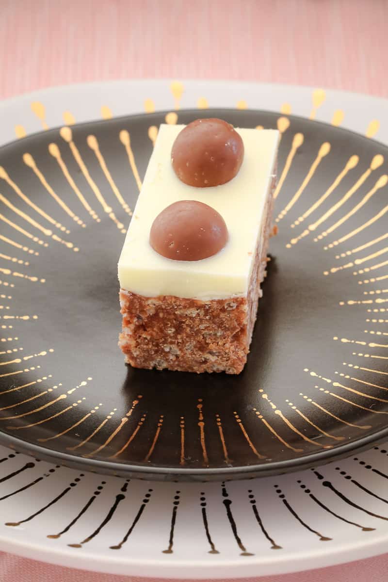 A piece of Malteser slice on a black plate, decorated with Maltesers and white chocolate icing.
