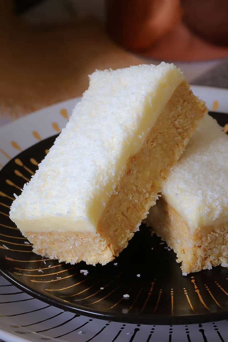 Two pieces of lemon coconut slice on a plate made with a crushed plain biscuit base, and a white lemon frosting on top.