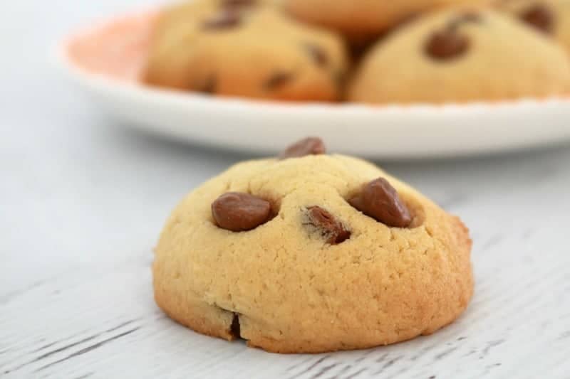 A close up of a chocolate chip cookie on a bench, in front of a plate filled with more cookies