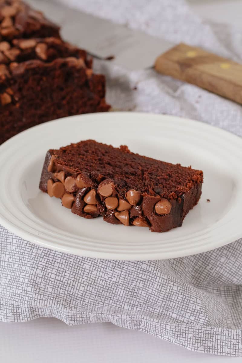 A slice of chocolate loaf topped with milk chocolate bits on a white plate.