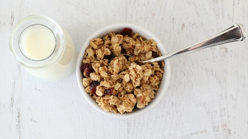 An overhead shot of granola in a small bowl with a spoon and a jar of milk beside it. 