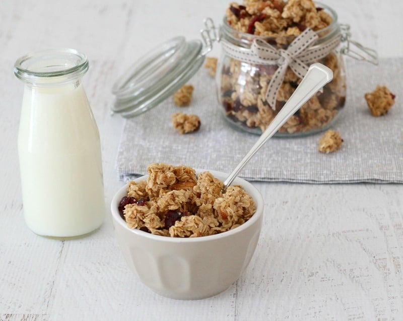 A small grey bowl filled with homemade granola with a bottle of milk beside and a jar of granola in the background.