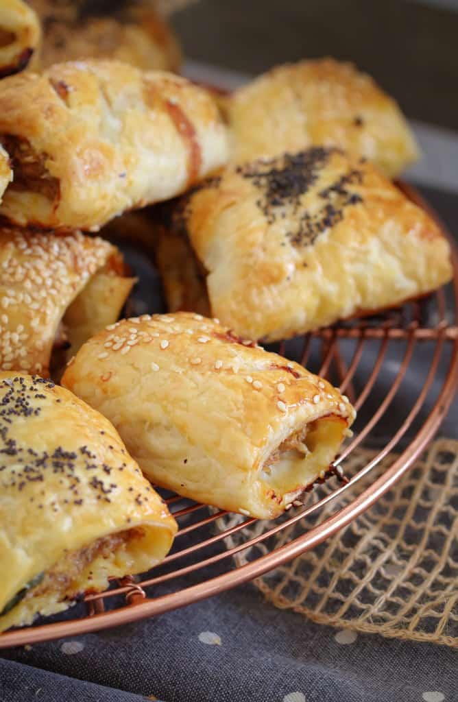 A bunch of golden sausage rolls, sprinkled with sesame seeds and poppy seeds on a wire copper rack.