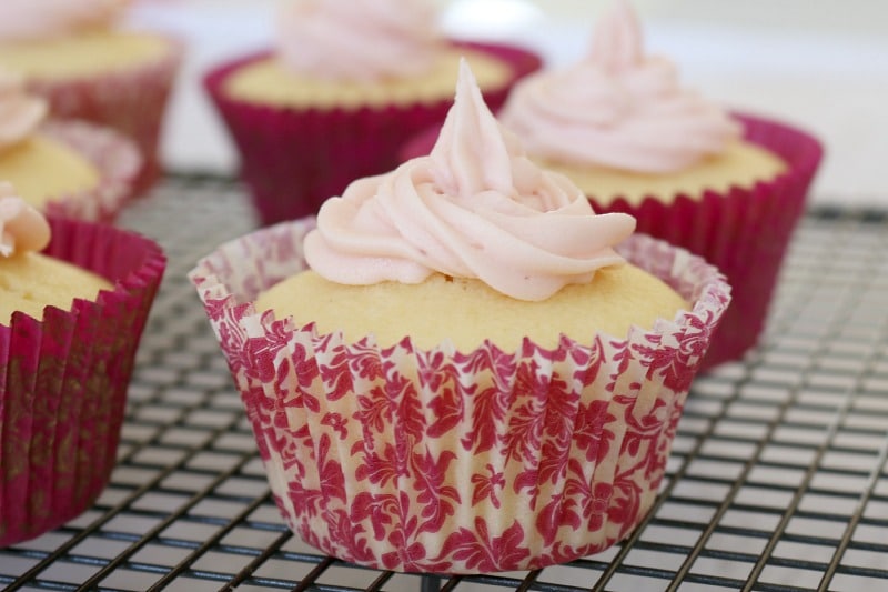 A close up of a lemon flavoured cupcake with white frosting in a pink floral cupcake case on a wire rack