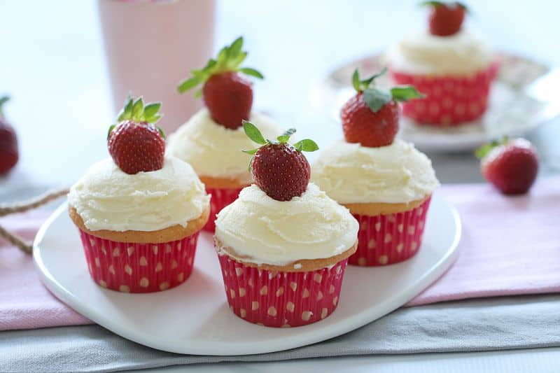 A white plate with four cupcakes topped with vanilla frosting and a fresh strawberry on each
