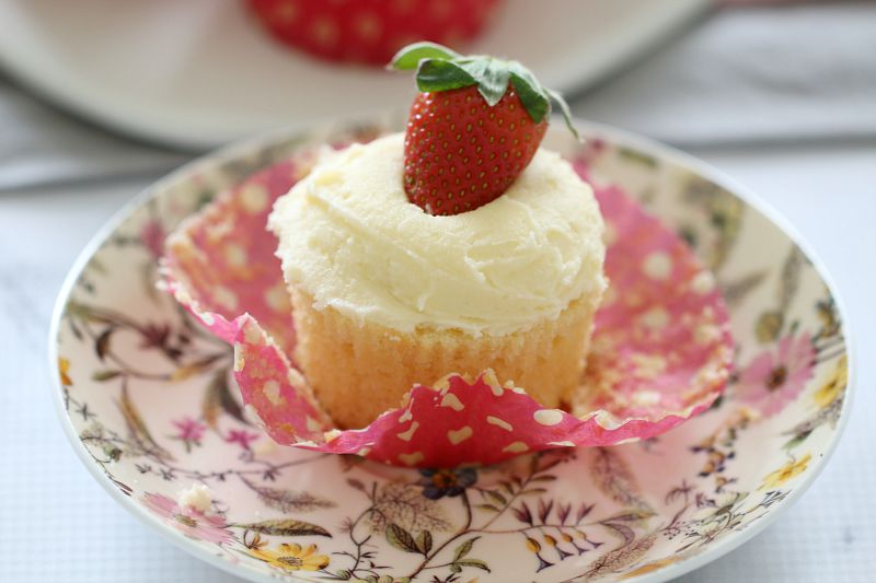 A vanilla cupcake served on a pretty floral plate, with vanilla frosting and a strawberry on top