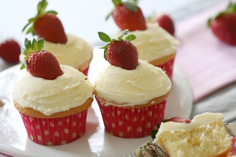 Cupcakes baked in pink and white cases, with white frosting and a fresh strawberry on top of each