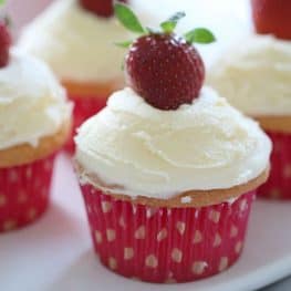 Vanilla cupcakes baked in pink and white cases, topped with white frosting and a fresh strawberry on top