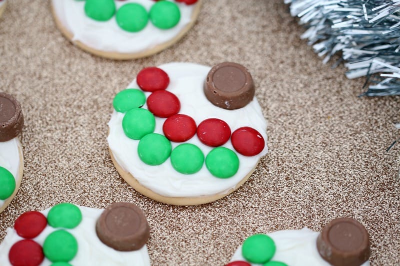 A close up of a biscuit topped with white frosting and decorated with red and green M&M\'s and a chocolate to resemble a Christmas bauble