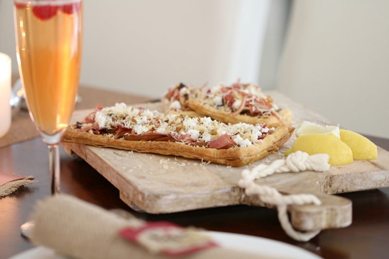 Two flat oblong pastry tarts topped with prosciutto, pear slices and goats cheese, served on a wooden board