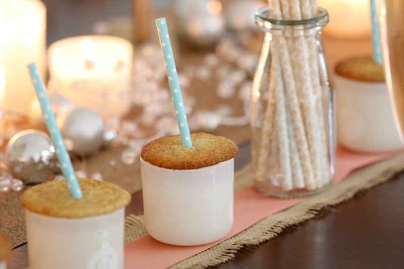 Spiced cookies on the top of white glass jars, with a straw through the middle of the biscuit, and Christmas decorations in the background