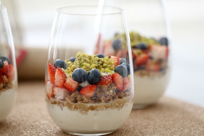 A close up of a glass with a layer of yoghurt in the base, then granola, mixed berries and topped with crushed pistachios