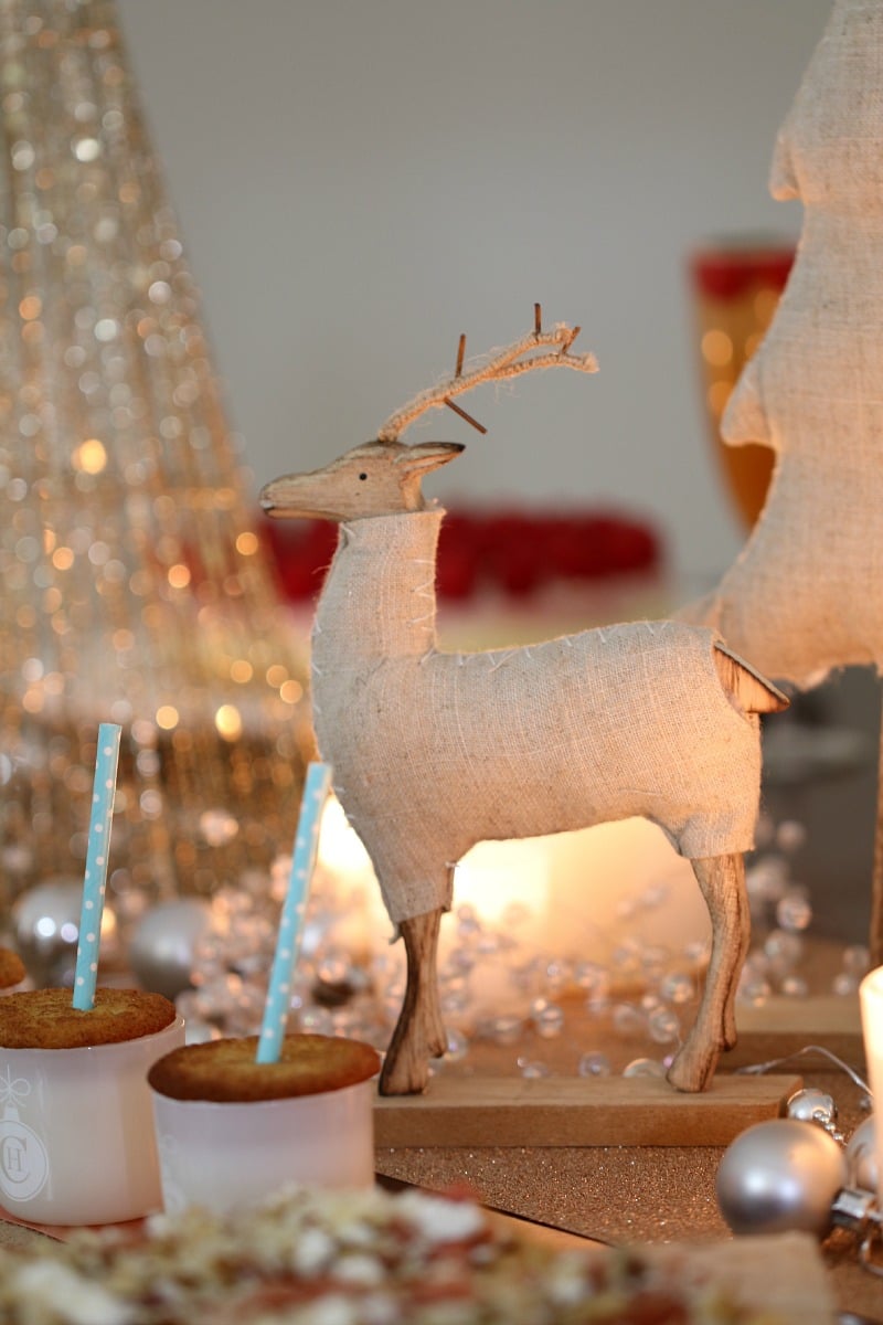 A close up of a wooden reindeer in front of white and gold Christmas decorations