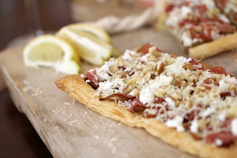 A close up of a flat pastry tart with prosciutto and goats cheese served on a board with wedges of lemon