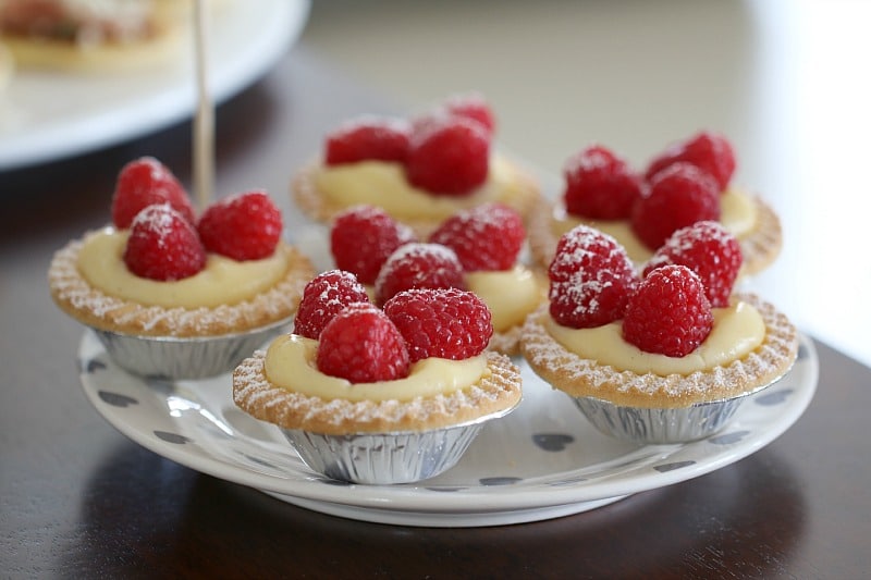 Individual custard tarts in foil cases, with fresh raspberries on top, and dusted with icing sugar
