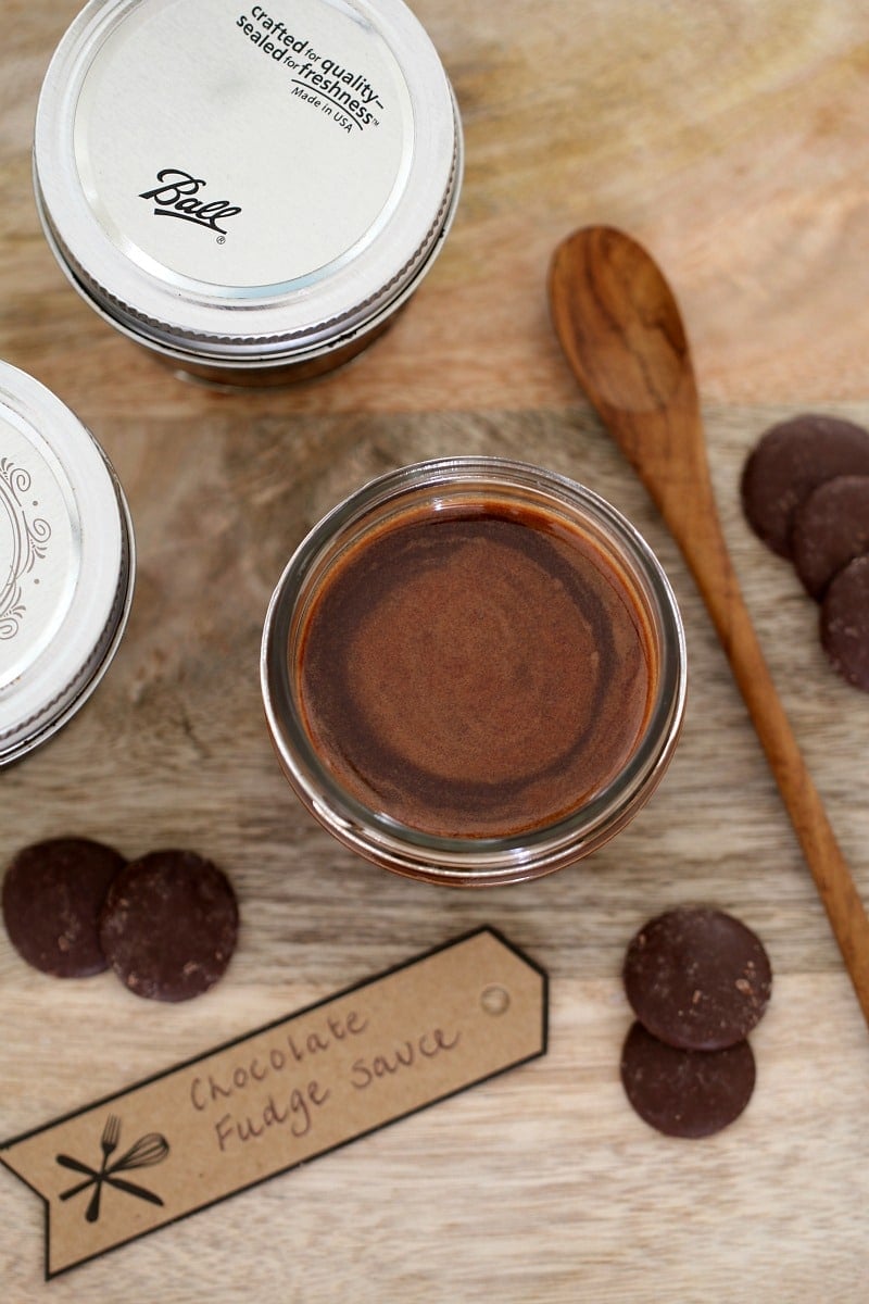 A glass jar filled with homemade chocolate sauce, with a label, a wooden spoon and chocolate buttons nearby