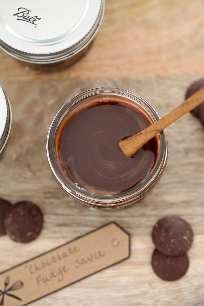 A wooden spoon in a glass jar filled with Chocolate Fudge Sauce, with a label and chocolate buttons nearby.