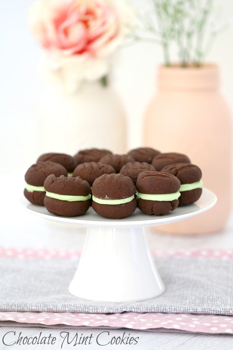 A white cake stand with a dozen chocolate cookies sandwiched together with mint icing