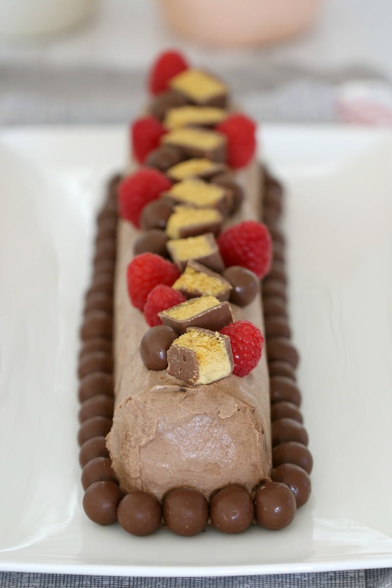 Maltesers around the base of a chocolate cream covered log, with chopped chocolate honeycomb and fresh raspberries on top
