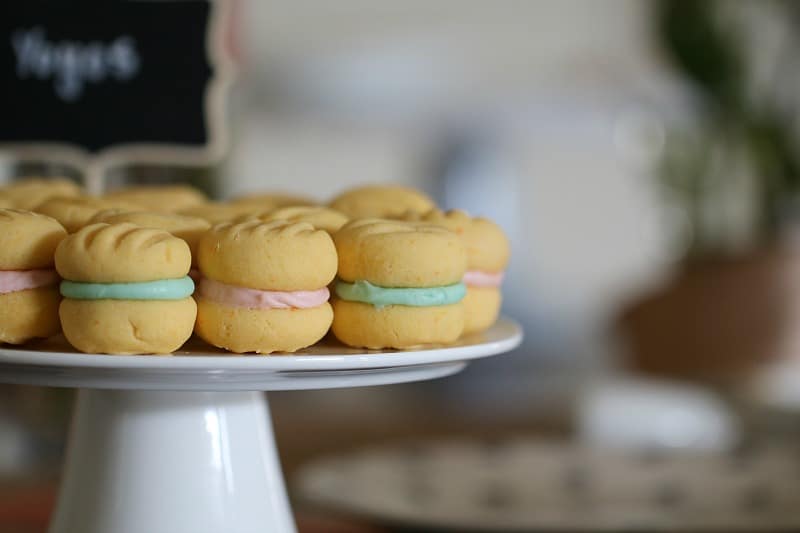 A white cake stand full of yo yo biscuits sandwiched together with pale blue and pink iced filling.