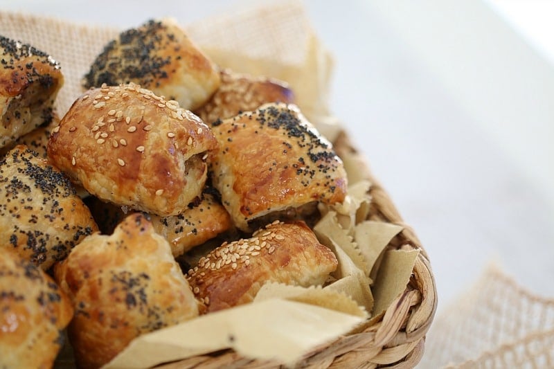 A close up of mini sausage rolls sprinkled with sesame and poppy seeds, piled in a basket