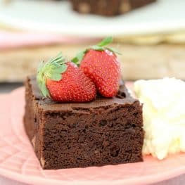 A square of chocolate brownie served on a plate with two fresh strawberries on top and a dollop of cream beside