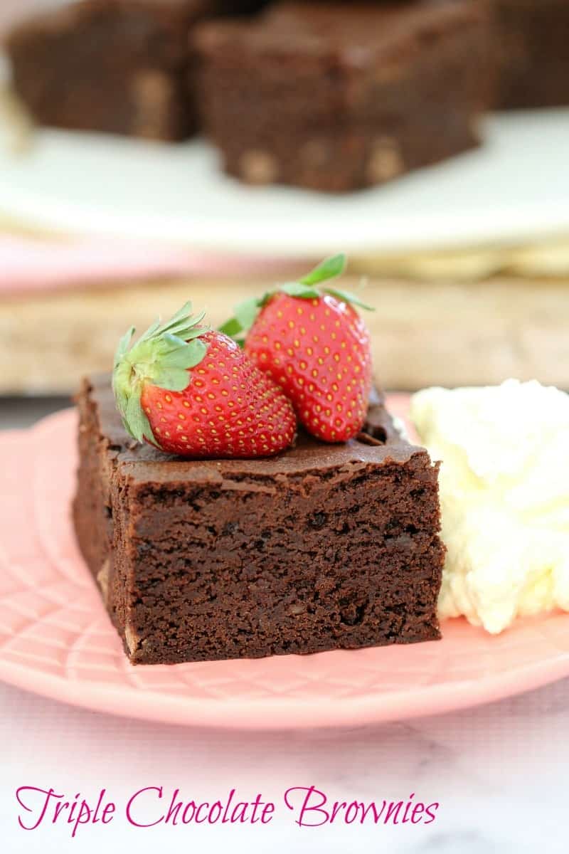 A square of chocolate brownie served on a plate with two fresh strawberries on top and a dollop of cream beside