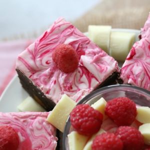 Swirly white chocolate and raspberry frosting on top of squares of brownie slice, with a bowl of fresh raspberries and white chocolate beside.