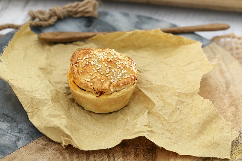 An individual meat pie baked golden with sesame seeds on top, sitting on a piece of brown paper