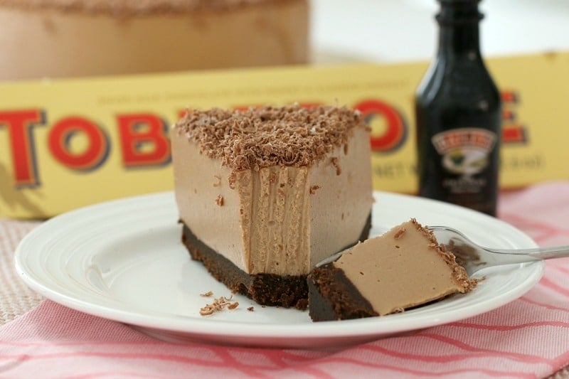 A slice of chocolate cheesecake on a white plate, in front of a Toblerone box and a bottle of Baileys.