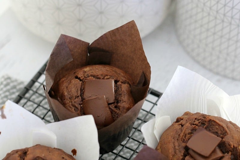 A close up of a baking paper wrapped chocolate muffin with chunks of chocolate on top, sitting on a wire tray