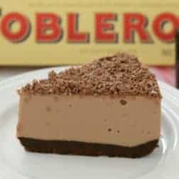 A piece of chocolate cheesecake on a white plate, decorated with grated chocolate in front of a box of Toblerone