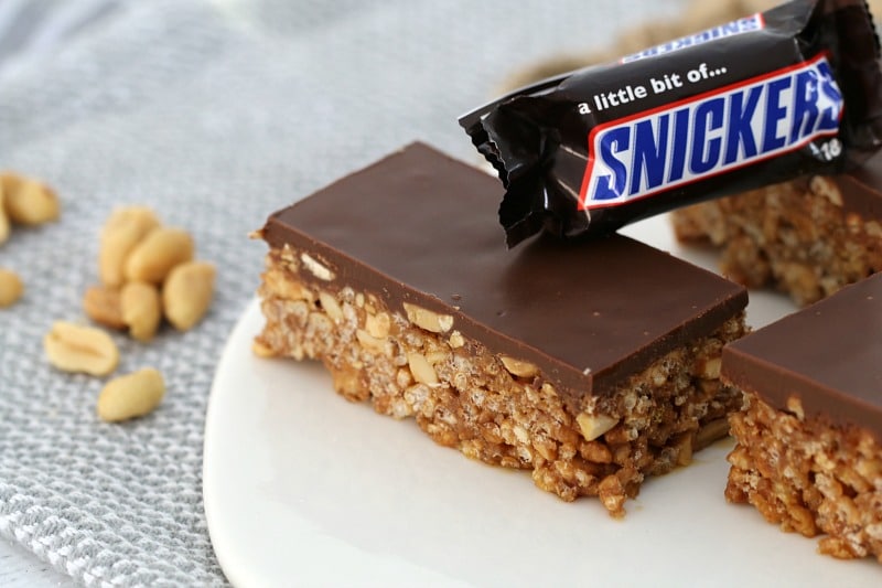 A piece of Snickers Slice on a white plate with a Snickers Bar resting on the slice