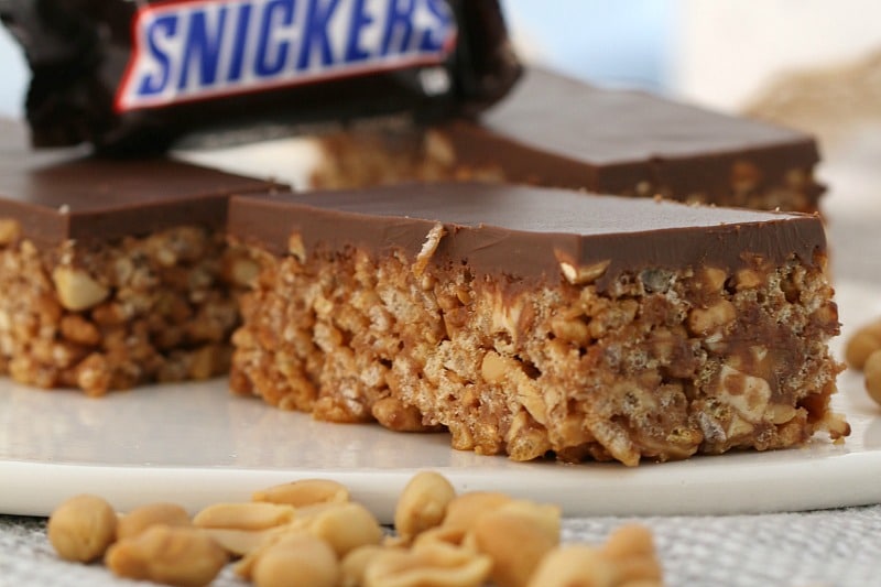 A close up of a piece of slice showing a filling made with rice bubbles, peanuts and Snickers Bars, and topped with milk chocolate