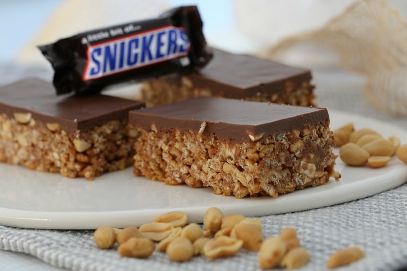 Pieces of Snickers Slice on a white board, with a mini Snickers bar on top and peanuts scattered beside