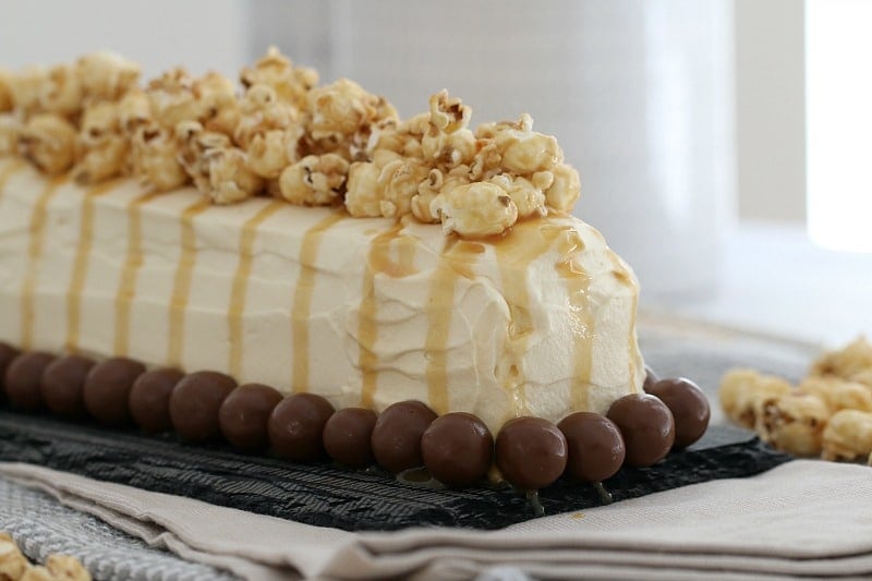 A cream covered log cake, with popcorn drizzled with caramel sauce on top, and Malteser balls around the base