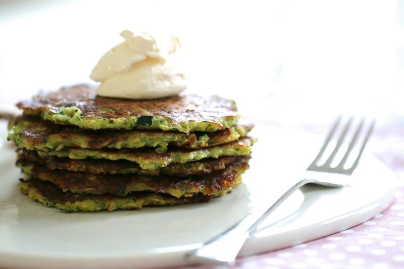 A stack of six thin vege fritters with a dollop of sour cream on top, served on a plate with a fork beside