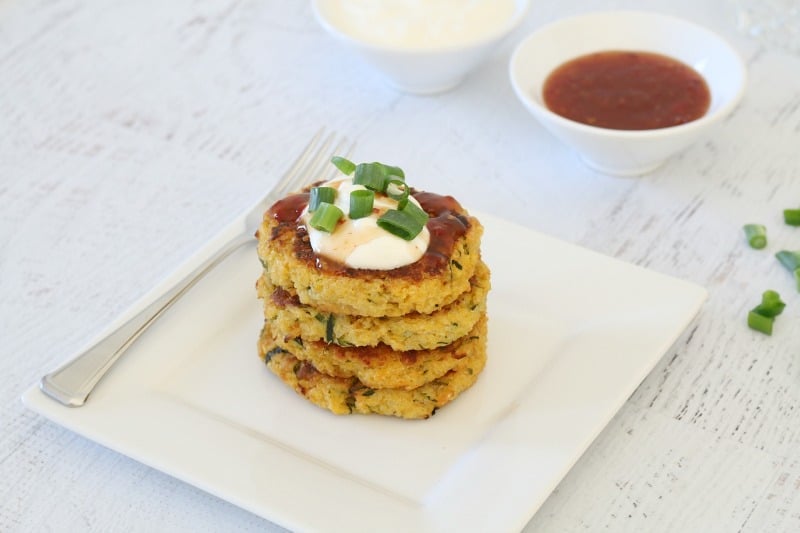 Four vege fritters stacked on a white plate, with a dollop of sour cream on top and a bowl of relish nearby