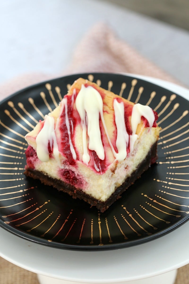 A cheesecake square made with raspberries and drizzled with white chocolate, served on a plate