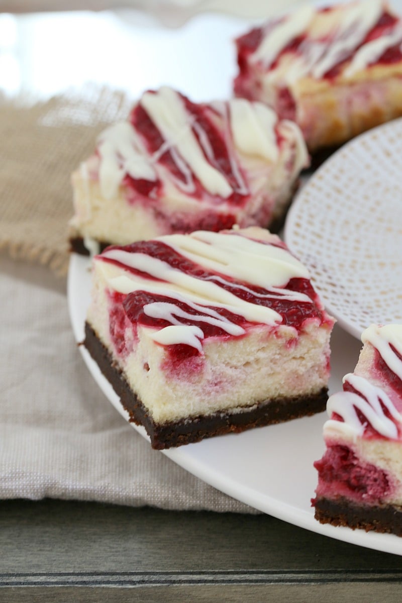 Squares of cheesecake with a chocolate base, a filling with raspberries and topped with white chocolate, served on a white plate 