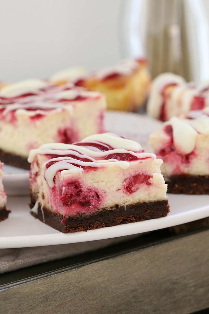 Cheesecake squares on a white plate showing a chocolate base, raspberries swirled through the filling and drizzled with white chocolate