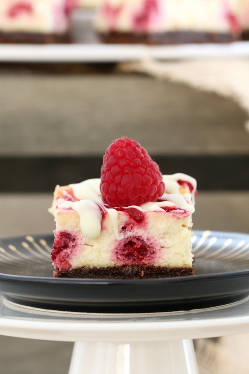 A square of cheesecake on a plate, with raspberries swirled through the white chocolate filling and topped with a fresh raspberry