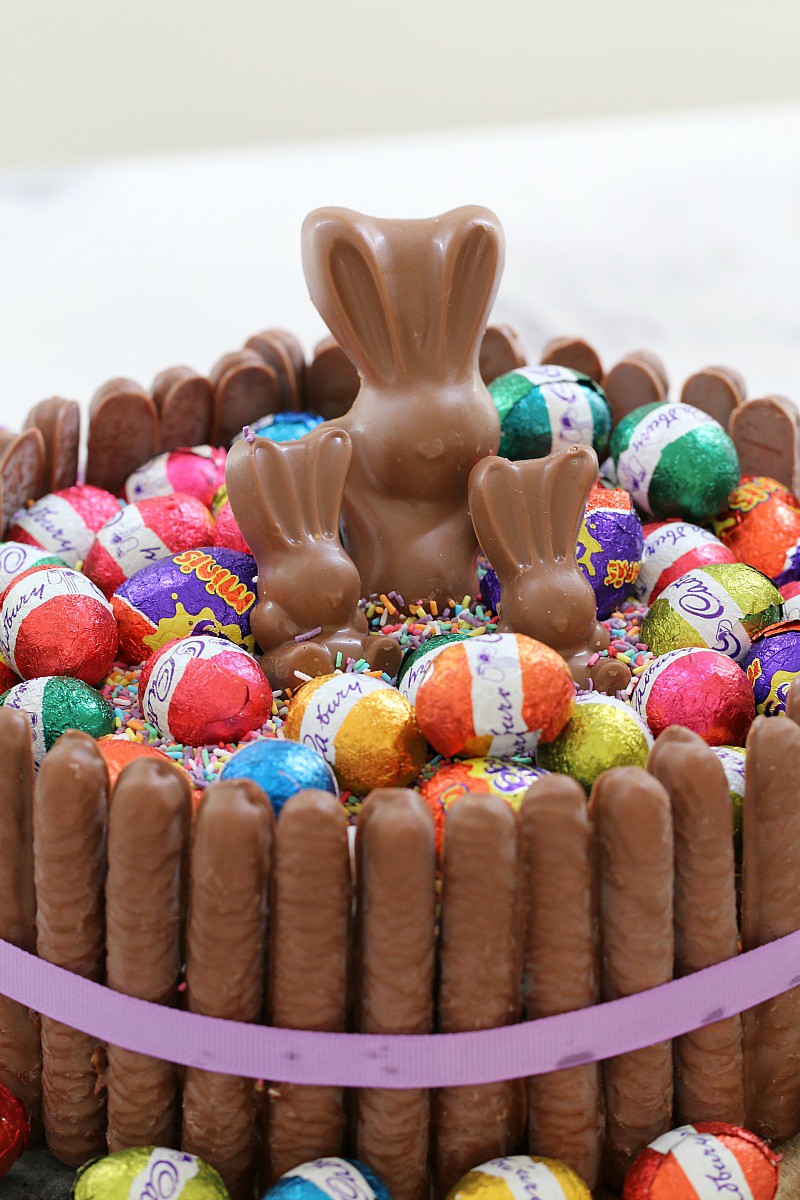 Wrapped mini Easter eggs, mini chocolate bunnies and colourful sprinkles on top of a round cake surrounded with chocolate finger biscuits