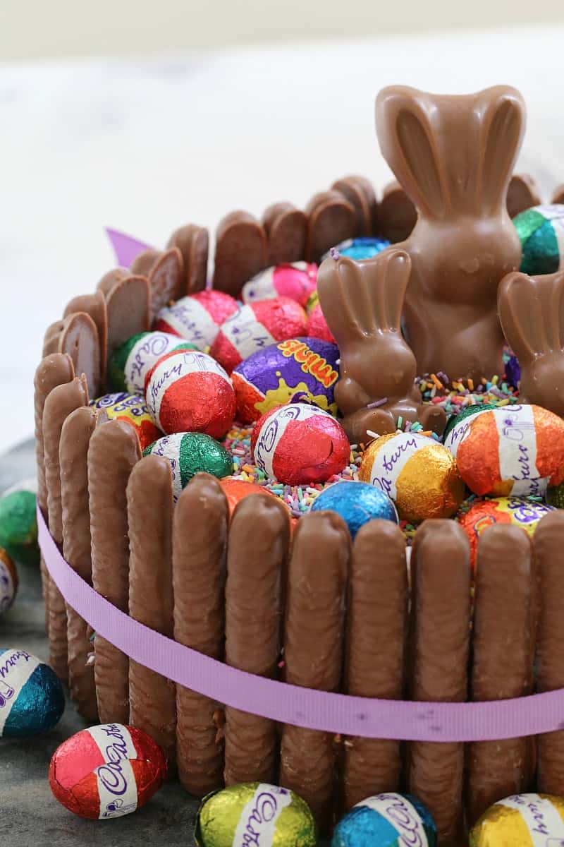 A close up of a cake surrounded with chocolate finger biscuits and topped with sprinkles, wrapped mini Easter eggs and mini chocolate bunnies