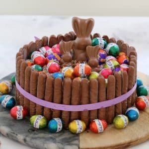 A round cake with wrapped mini Easter eggs and chocolate bunnies on top, with chocolate finger biscuits around the sides, and more mini Easter eggs