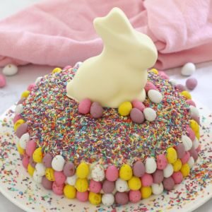 A round cake decorated with pretty pastel sprinkles, mini pastel Easter eggs and a white chocolate bunny on top
