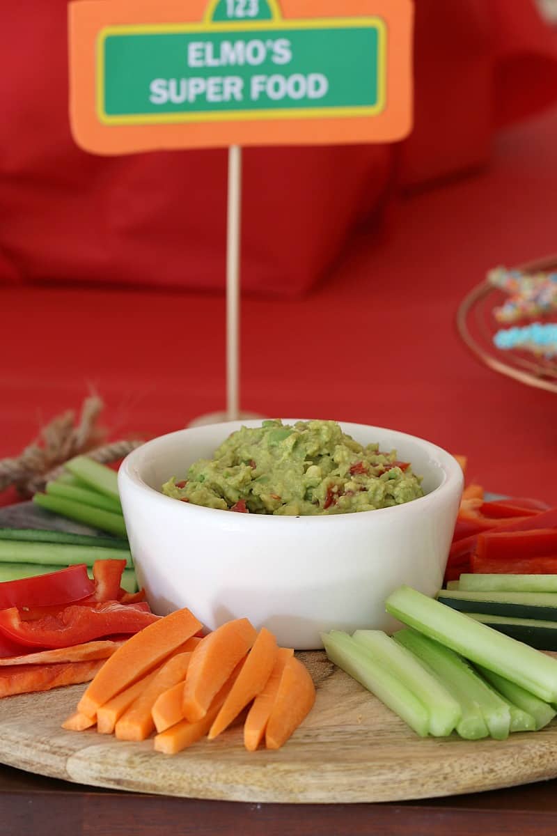 A small white bowl of avocado dip surrounded by celery, carrot and capsicum sticks for dipping
