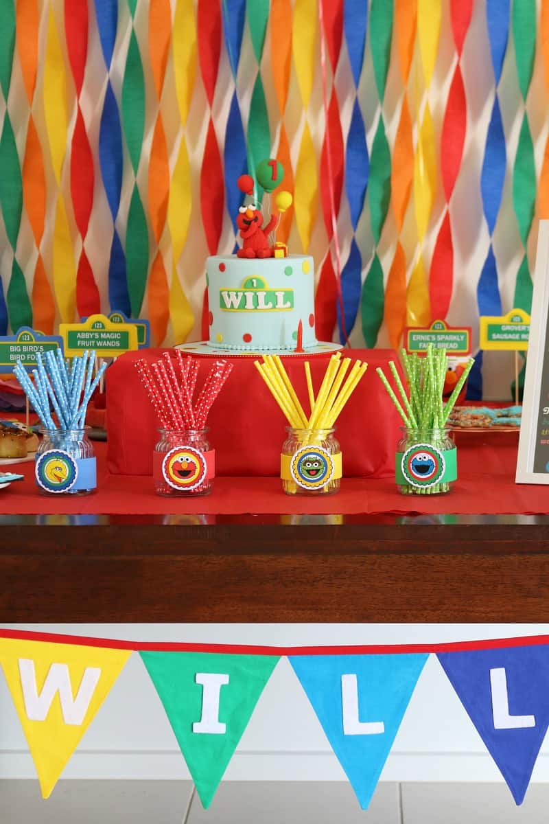 Coloured straws in jars in front of a Sesame Street decorated cake with Cookie Monster on top