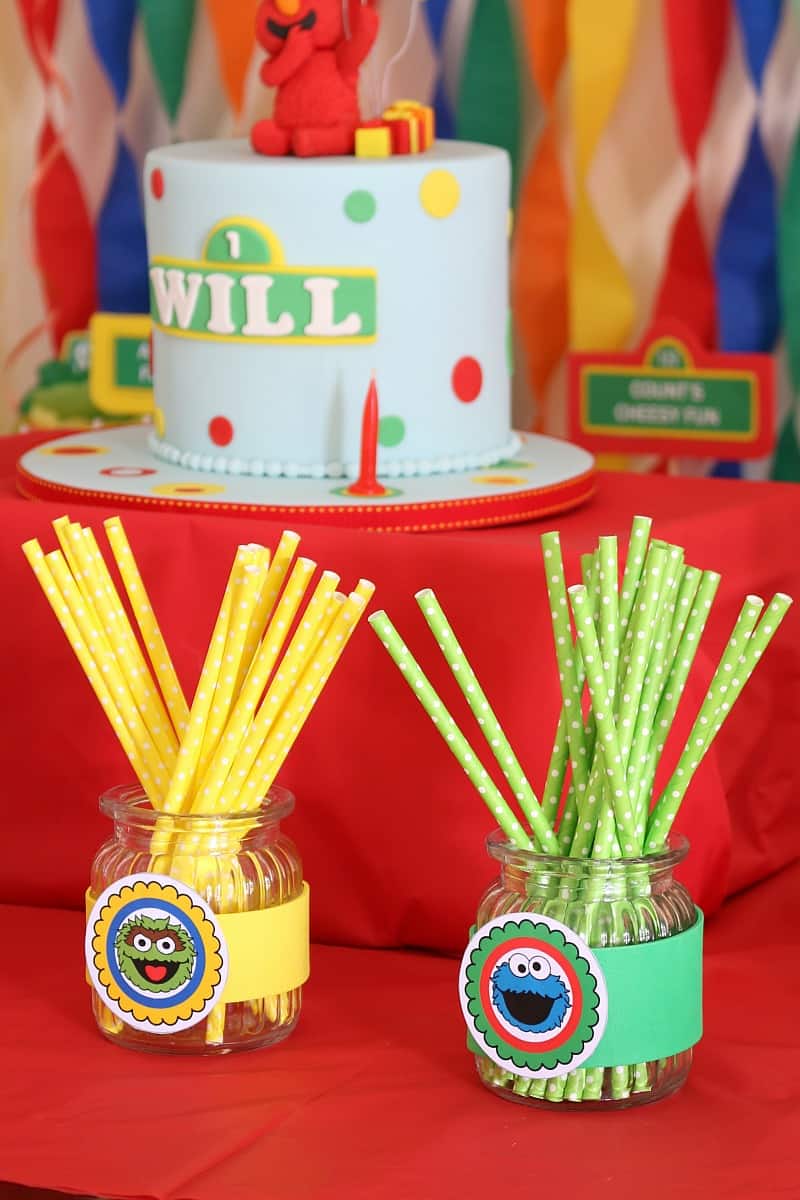 Colourful straws in jars in front of a pale blue Sesame Street decorated cake
