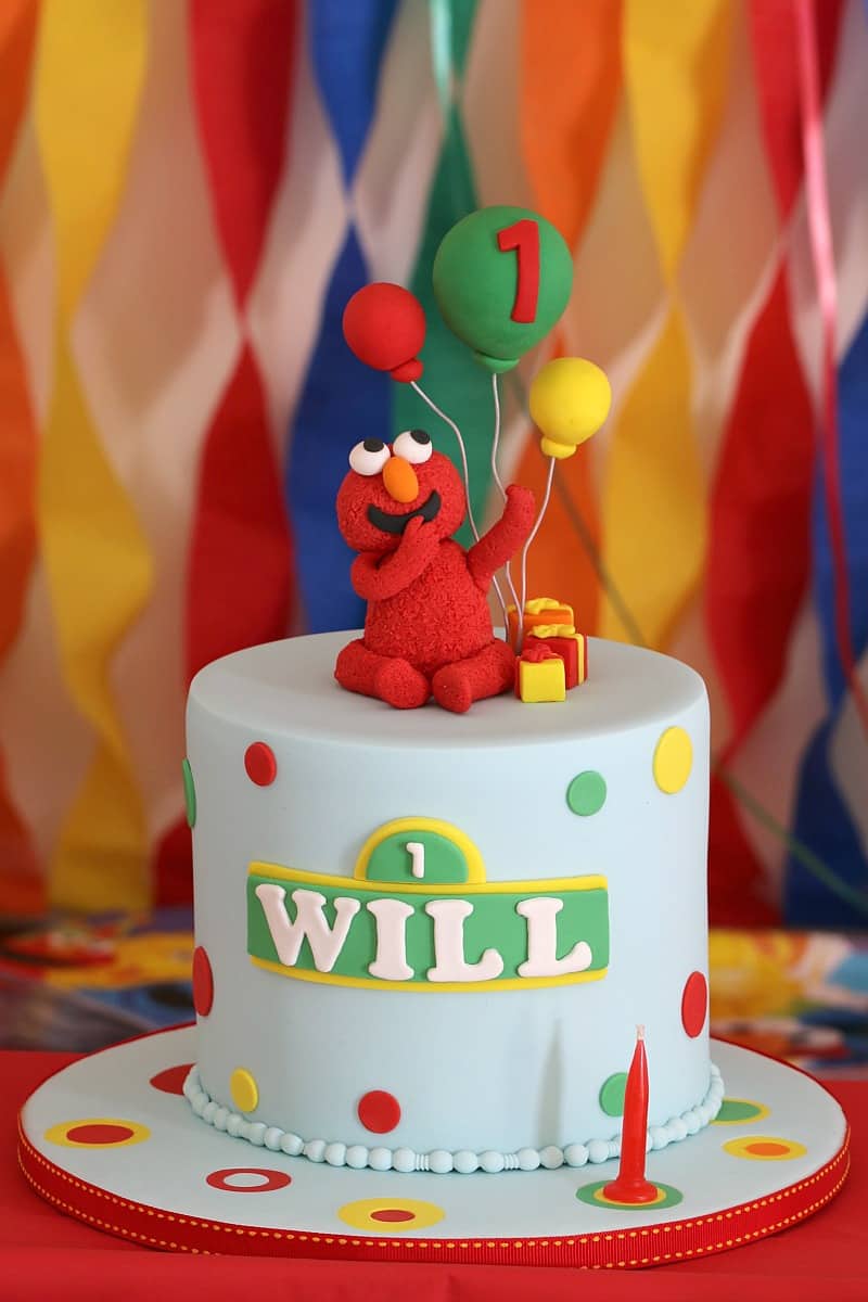 A tall round birthday cake, iced pale blue, with an edible Cookie Monster holding balloons sitting on top